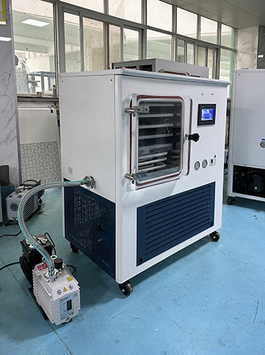 Left perspective real shot of a white laboratory freeze dryer in a factory workshop, connected via hose to an external vacuum pump.
